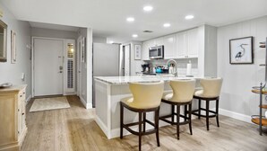 A modern kitchen with white cabinetry, a central island with bar stools, a microwave, and a coffee maker. - Surf Court 62 - Forest Beach 1st Floor Flat (Hilton Head Island)