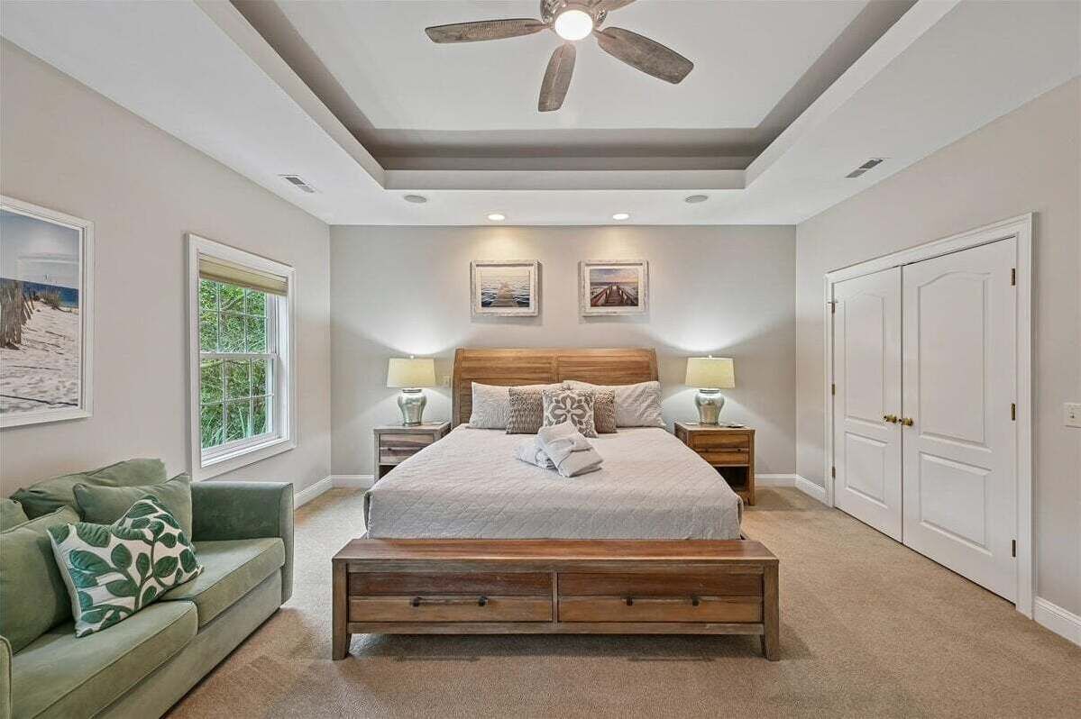 A bedroom with a bed, a ceiling fan, a nightstand, a bedside table, a green couch, and a window with a view of trees.