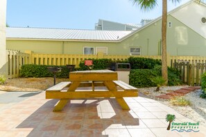 A picnic table in a courtyard with a yellow fence, a small palm tree, and a building with a white roof. - Surfside Sandcastle-Sunbird-Beachfront PCB, King Suite-Book Summer Vacay Now! (Panama City Beach)