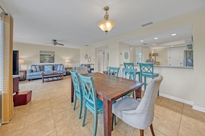 A spacious dining area with a wooden table and turquoise chairs, a living room with a sofa and TV, and a kitchen with a bar.