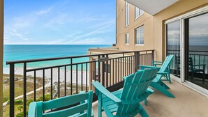 Uma varanda com cadeiras Adirondack turquesa, vista para a praia e um edifício com janelas grandes.