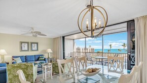 A spacious living room with a balcony overlooking the ocean, featuring a glass dining table, blue sofa, and a chandelier.