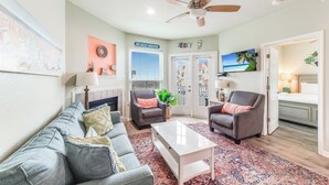 A living room with a grey sofa, two armchairs, a coffee table, a ceiling fan, a TV, and a door labeled "BEACH HOUSE".