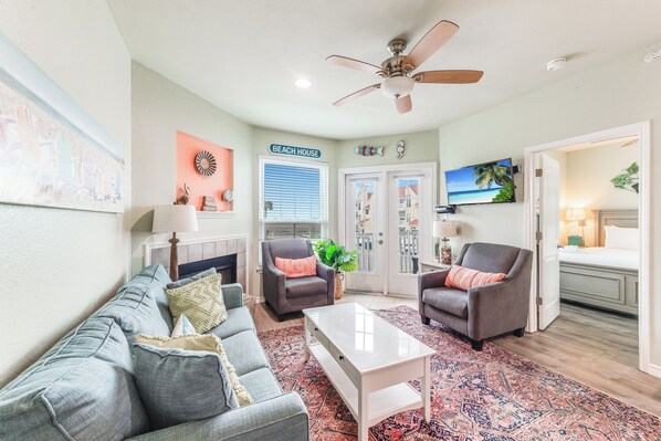 Ein Wohnzimmer mit grauem Sofa, zwei Sesseln, einem Couchtisch, einem Deckenventilator, einem Fernseher und einer Tür mit der Aufschrift "BEACH HOUSE".