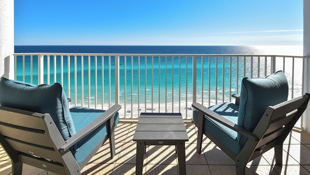 A balcony with two chairs and a table, overlooking a beach with clear turquoise waters and a white railing.