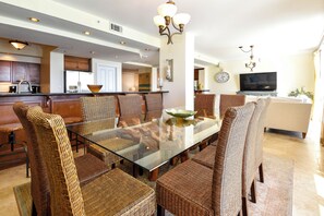 Een ruime eethoek met een tafel met glazen blad, omringd door rieten stoelen, een keuken op de achtergrond en een zithoek met een televisie en klok.