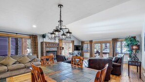 A spacious living area with a large wooden dining table, multiple seating options including a sofa and chairs, a fireplace, and large windows allowing natural light. - Downtown Gem - Ski Resort Views, Private Hot Tub, Dog-Friendly MainD (Breckenridge)