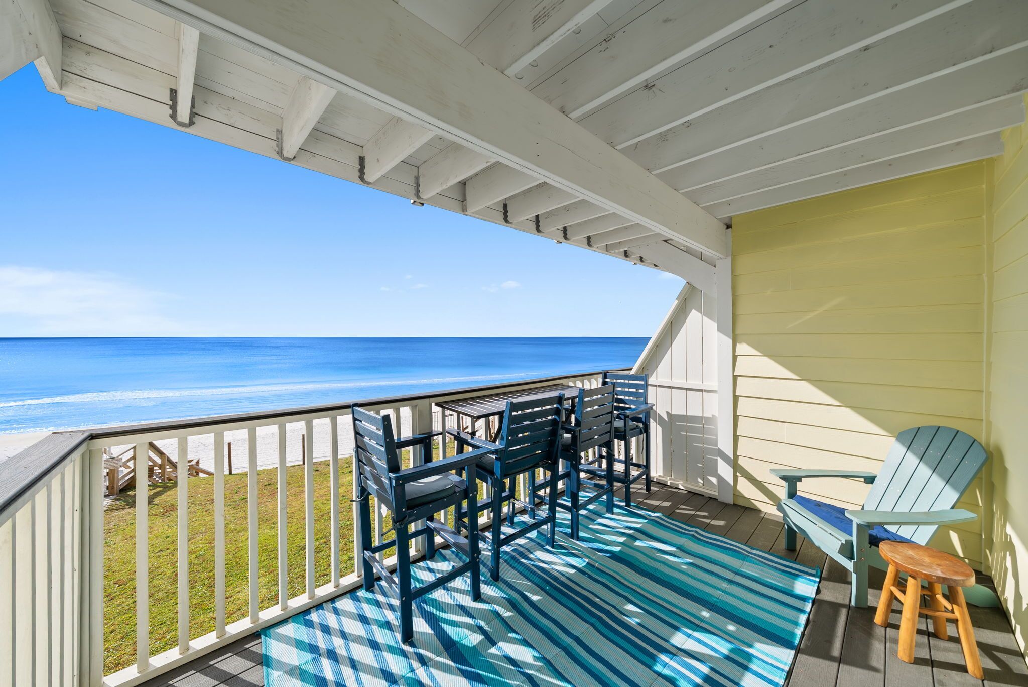 A covered deck with a view of the ocean, featuring a blue and white striped rug, a wooden chair, and a small stool.
