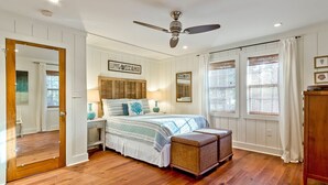 A bedroom with a wooden floor, a bed with a striped bedspread, a ceiling fan, and a window with curtains. - Just Beachy | Pet Friendly, Pool, Fenced Yard! (Tybee Island)