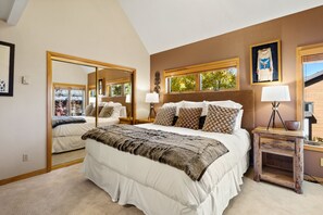 A bedroom with a large bed, a nightstand, a mirror, and a window with a view of trees.