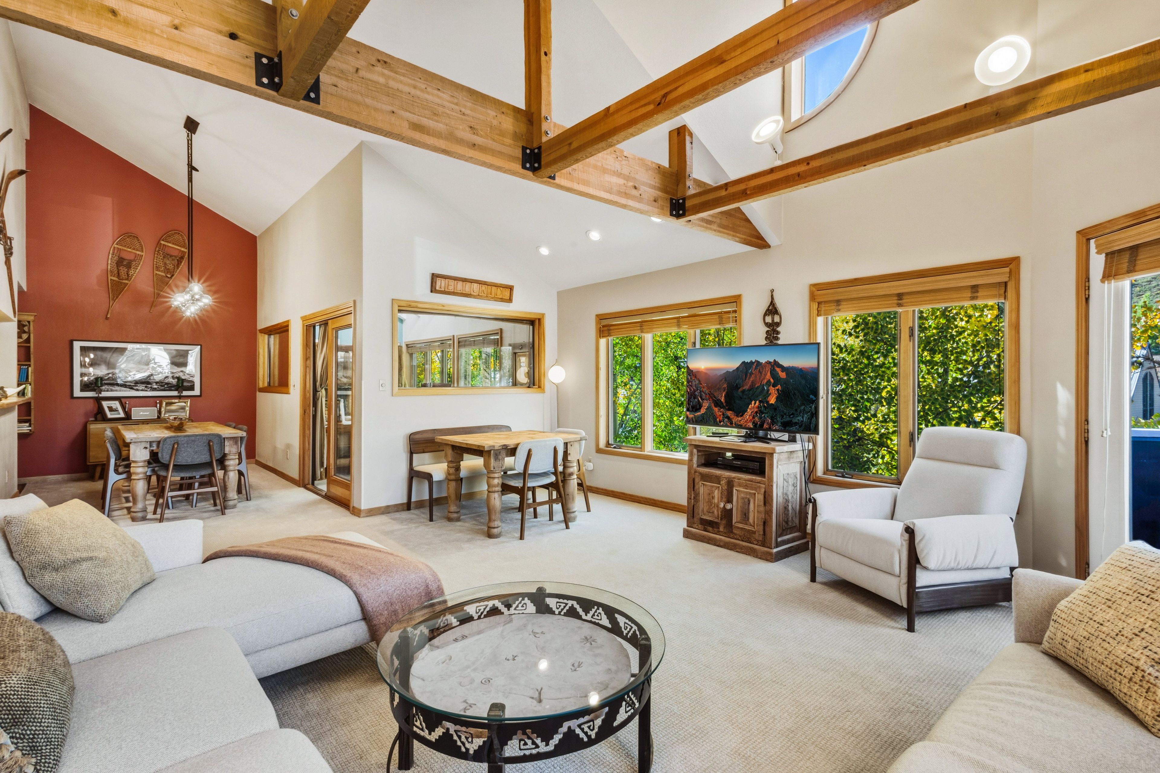 A spacious living room with a high ceiling, wooden beams, a large TV, and a dining area.