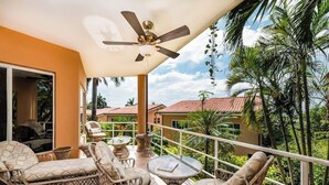 A balcony with wicker furniture, a ceiling fan, and a view of palm trees and houses.
