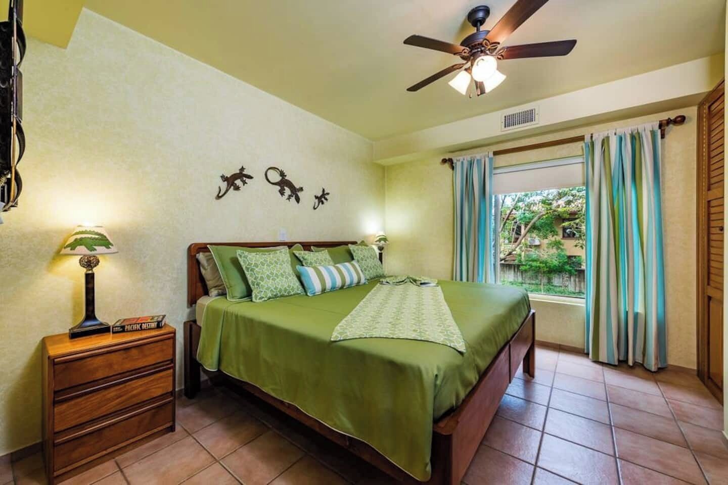 Une chambre à coucher comprenant un lit, une table de chevet, une lampe, un ventilateur de plafond et une fenêtre avec des rideaux.