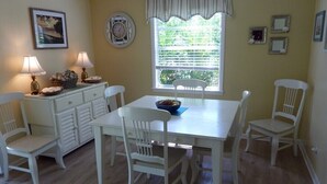 Un comedor con una mesa y sillas blancas, una ventana con persianas, un reloj en la pared y un cuadro colgado sobre la mesa.