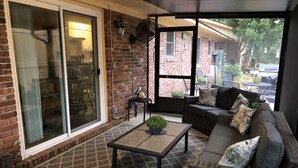 Un patio cubierto con pared de ladrillo, puerta corrediza de vidrio, mobiliario de mimbre y una pequeña mesa con una planta.