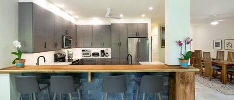 A modern kitchen with dark cabinets, a wooden island with bar stools, a stainless steel refrigerator, and a ceiling fan.