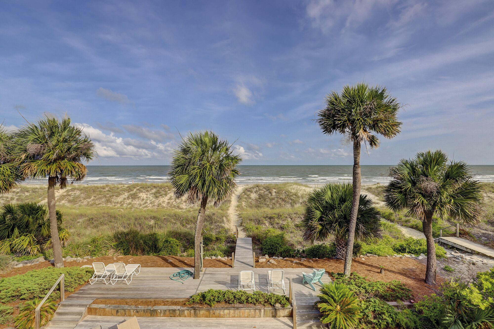 A beach view with palm trees, a wooden deck with lounge chairs, and a clear sky.