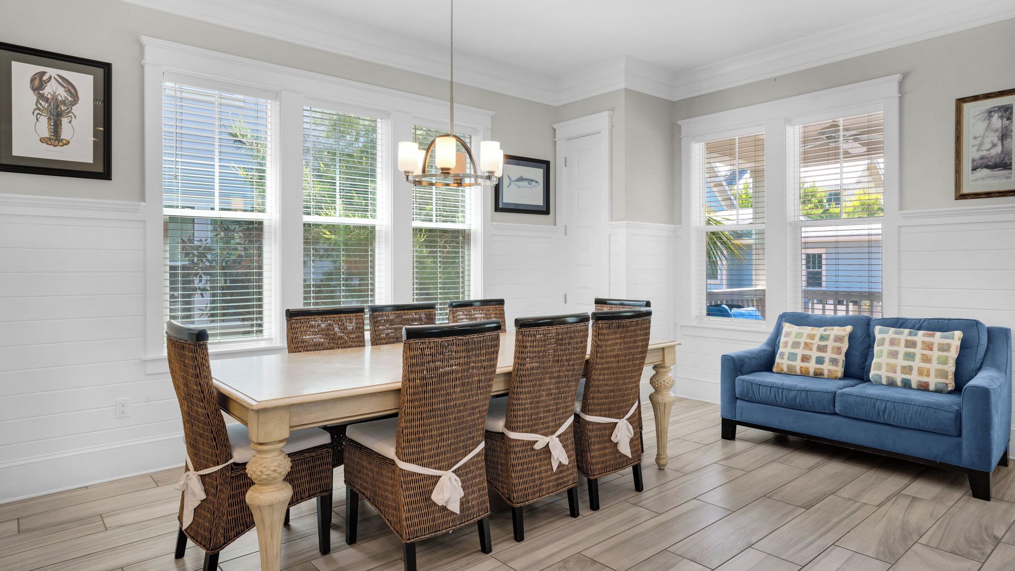 A dining area with a wooden table and wicker chairs, a blue sofa, and large windows with blinds.
