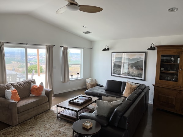 A spacious living room with a large window, a ceiling fan, a wooden cabinet, and a framed landscape painting. - Vineyard Views + Fire Pit | 1 Min to Wine Trail | Top of the Hill (San Miguel)