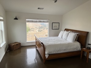 A bedroom with a wooden bed, a window with a view of a landscape, a small bedside table, and a wall-mounted light fixture. - Vineyard Views + Fire Pit | 1 Min to Wine Trail | Top of the Hill (San Miguel)
