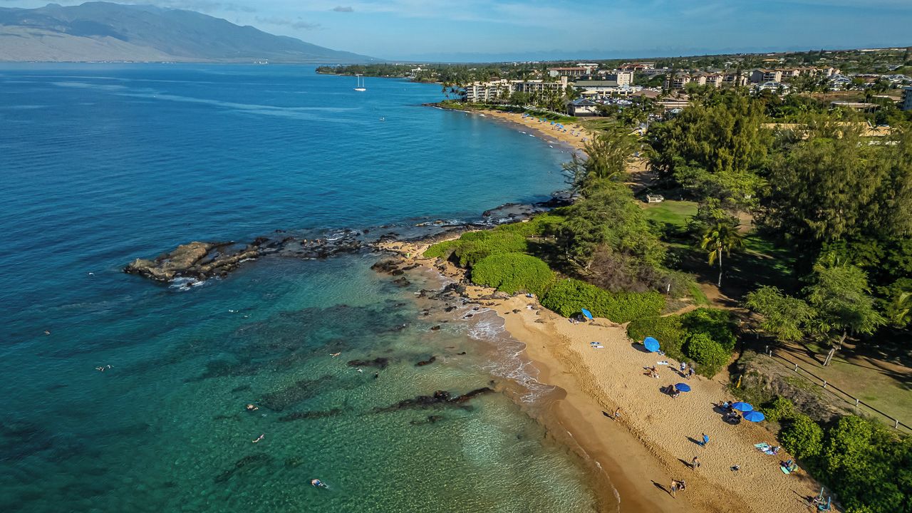 Ein Küstenstrand mit klarem Wasser, Sandstrand und Wohnhäusern im Hintergrund.