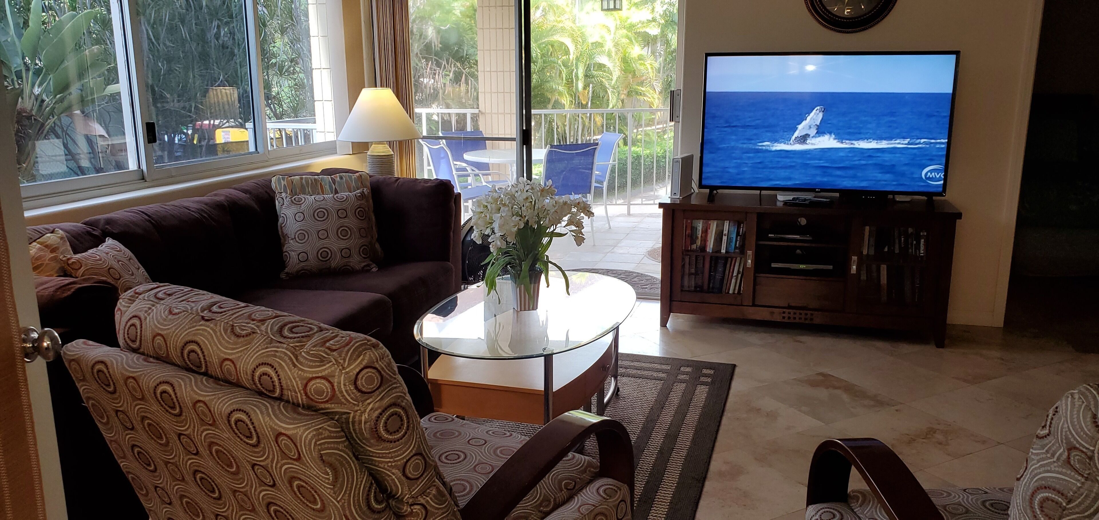 Una sala de estar con un sofá, sillones estampados, una mesa de centro con un jarrón, un televisor sobre un mueble bajo y una estantería.