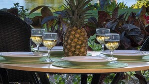 A table set with two glasses of white wine, a pineapple, and a plate, overlooking a body of water with trees and a clear sky.