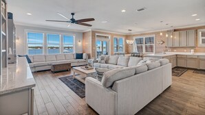 A spacious living room with a large grey sectional sofa, a dining area, and a kitchen with wooden cabinets and a white countertop. - Good Time Charlie by GSVR | Lagoonfront Home w/ Pool, Firepit & Beach Access + FREE Activities/Golf (GULF SHORES)
