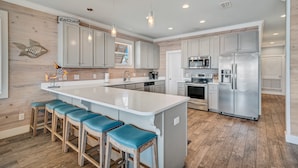 A modern kitchen with a central island, bar stools, stainless steel appliances, and wooden flooring. - Good Time Charlie by GSVR | Lagoonfront Home w/ Pool, Firepit & Beach Access + FREE Activities/Golf (GULF SHORES)