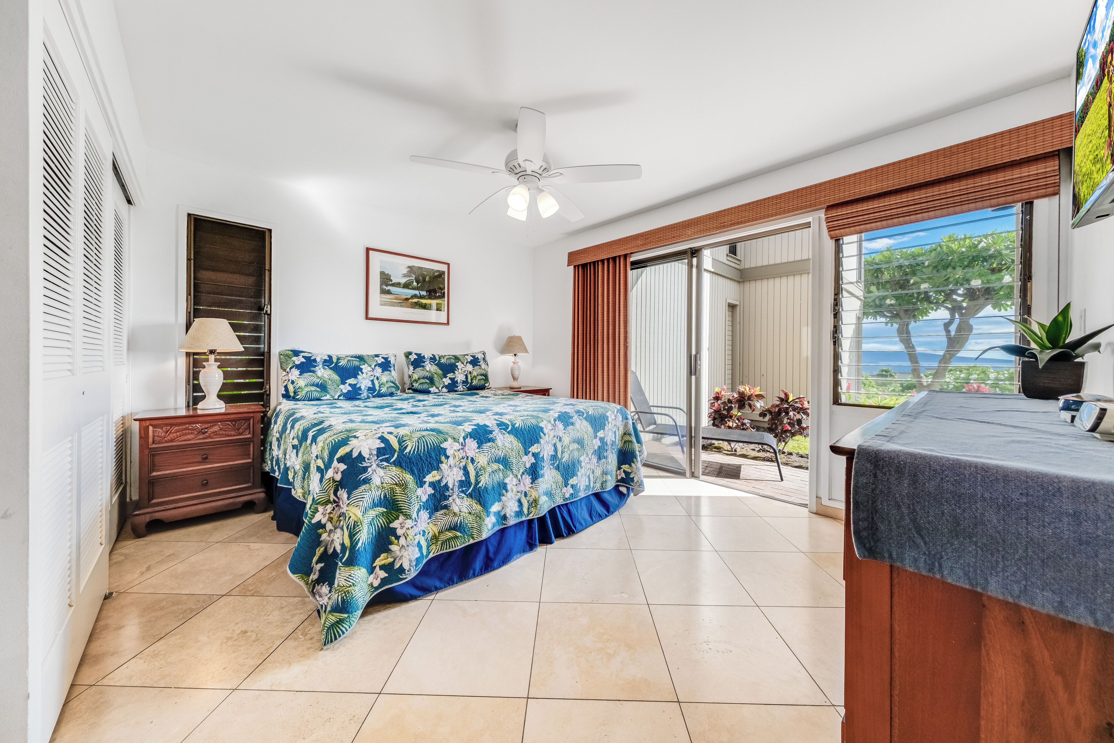 Une chambre à coucher comprenant un lit, une table de chevet, une lampe, un ventilateur de plafond, une porte-fenêtre coulissante donnant sur un balcon et un tableau encadré accroché au mur.