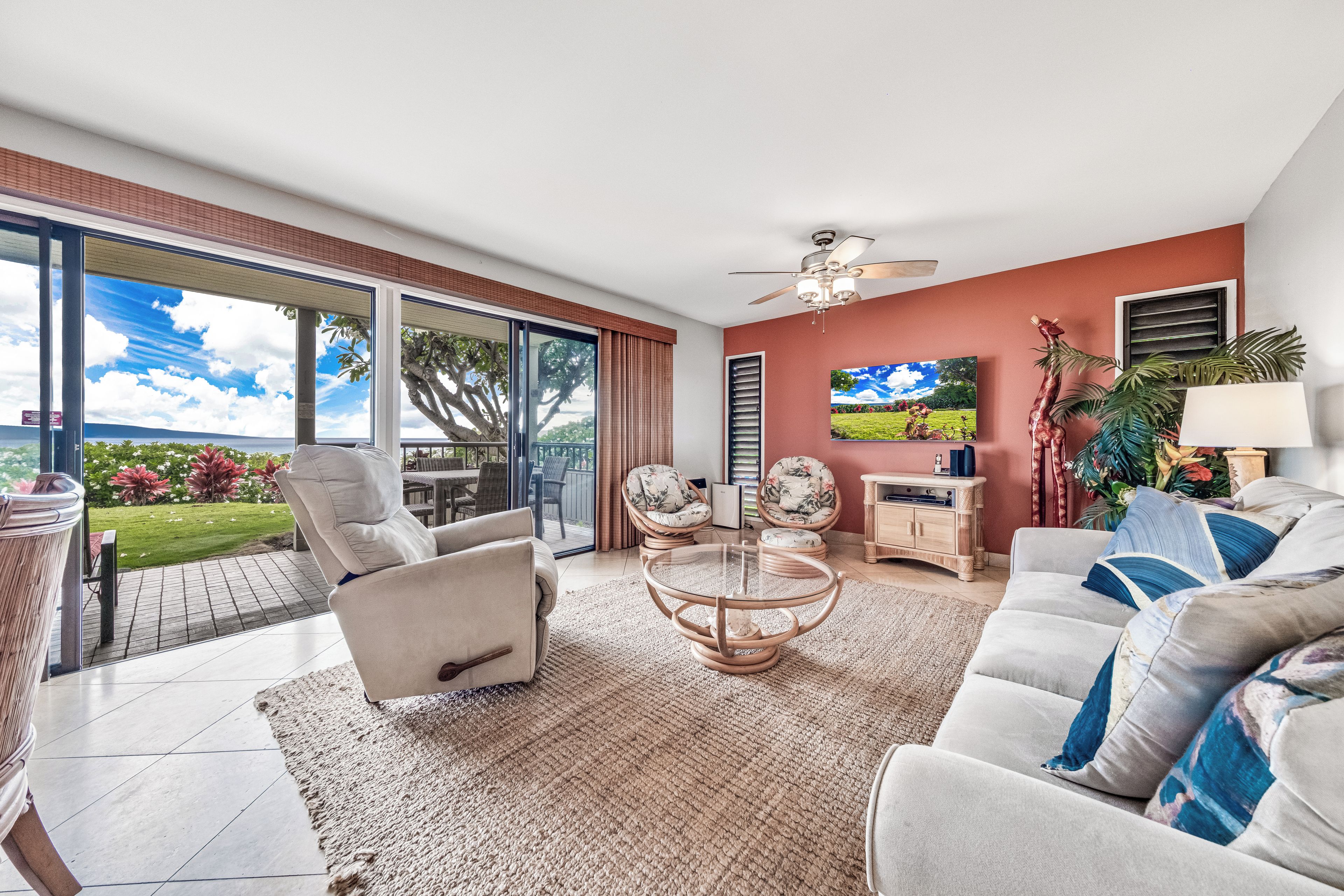 A spacious living room with a large sliding glass door leading to a patio, a ceiling fan, a flat-screen TV, and a dining area with wicker furniture.