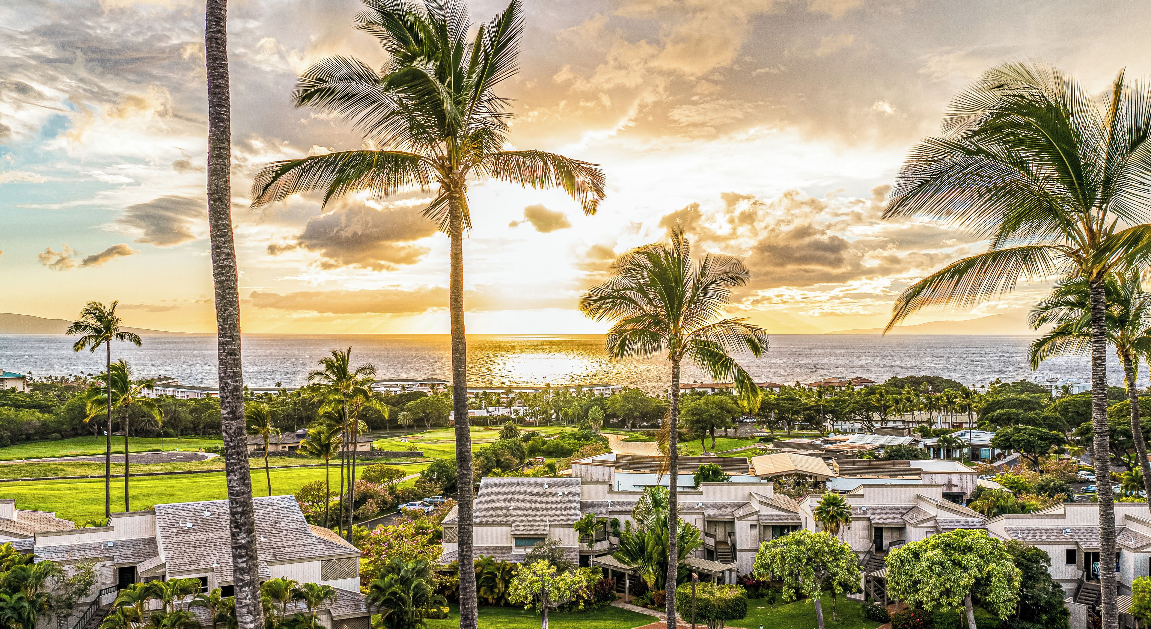 A coastal resort with multiple-story buildings, lush greenery, and a view of the ocean at sunset.