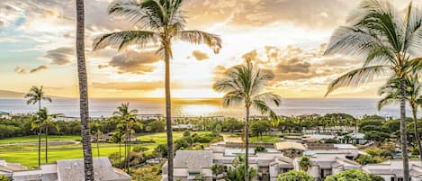 A coastal resort with multiple-story buildings, lush greenery, and a view of the ocean at sunset.