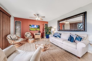 Un salon avec un canapé blanc, deux fauteuils, une table basse, un ventilateur de plafond, un grand miroir et un tableau accroché au mur.