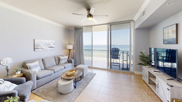 A modern living room with a sofa, coffee table, and TV, leading to a balcony with a view of the beach.