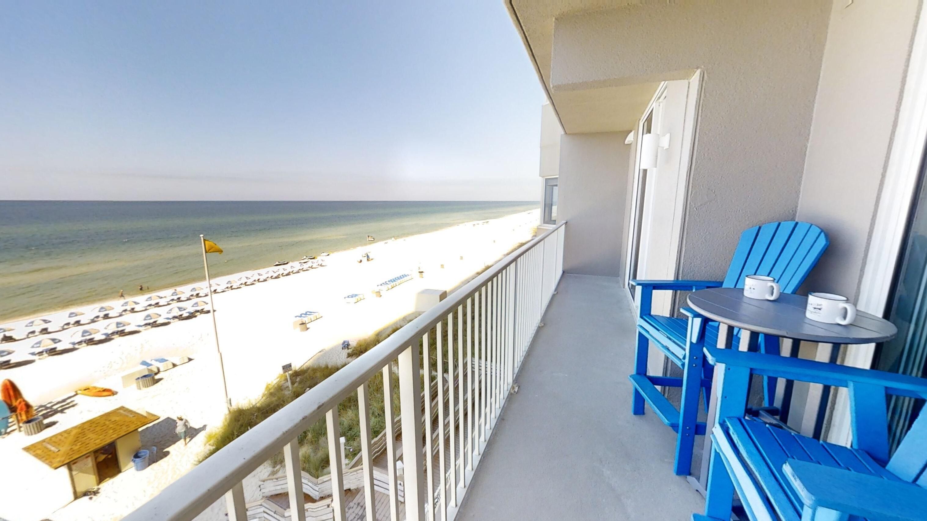A balcony with a view of the beach, blue chairs, and a table with cups.