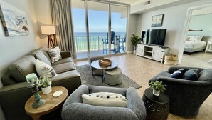 A modern living room with a grey sofa, a grey armchair, a round coffee table, and a TV stand. There is a balcony with a view of the ocean. - Tidewater 309 - Beachfront Bliss with Breathtaking Gulf Views (Panama City Beach)