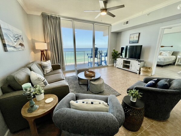 A modern living room with a grey sofa, a grey armchair, a round coffee table, and a TV stand. There is a balcony with a view of the ocean.