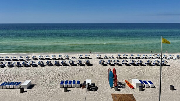 A beach with rows of sun loungers and umbrellas, a flag, and a sign indicating a banana boat service. - Tidewater 309 - Beachfront Bliss with Breathtaking Gulf Views (Panama City Beach)