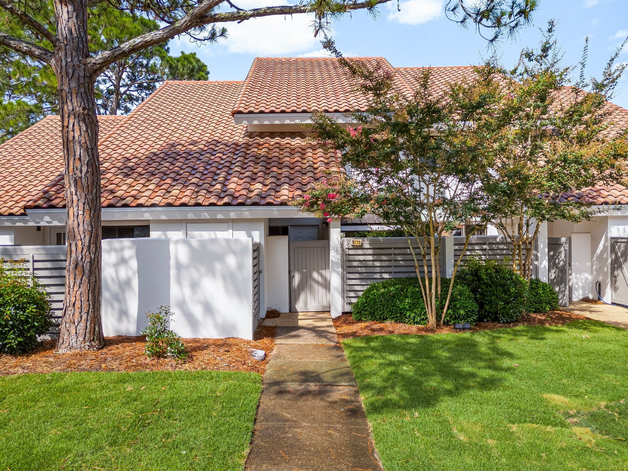 A two-story house with a red-tiled roof, a front door, and a small front yard with a tree and shrubs.