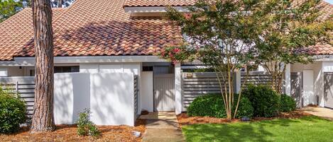 A two-story house with a red-tiled roof, a front door, and a small front yard with a tree and shrubs.