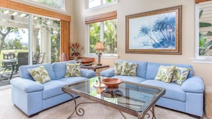 A spacious living room with a blue sofa set, a glass coffee table, and a large framed painting on the wall. - Comfortable Townhome in Waikoloa Resort - Walk to Shops! (Waikoloa)
