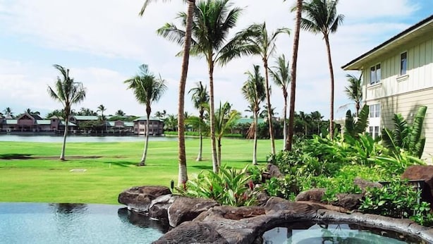 A pool with natural rock formations, surrounded by lush greenery and palm trees, with a view of a golf course and resort buildings in the background. - Comfortable Townhome in Waikoloa Resort - Walk to Shops! (Waikoloa)