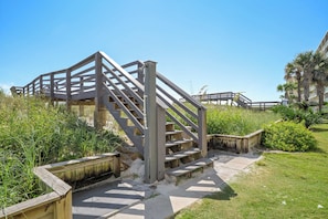A wooden staircase leading to a beach area with a railing, surrounded by greenery and a clear sky.