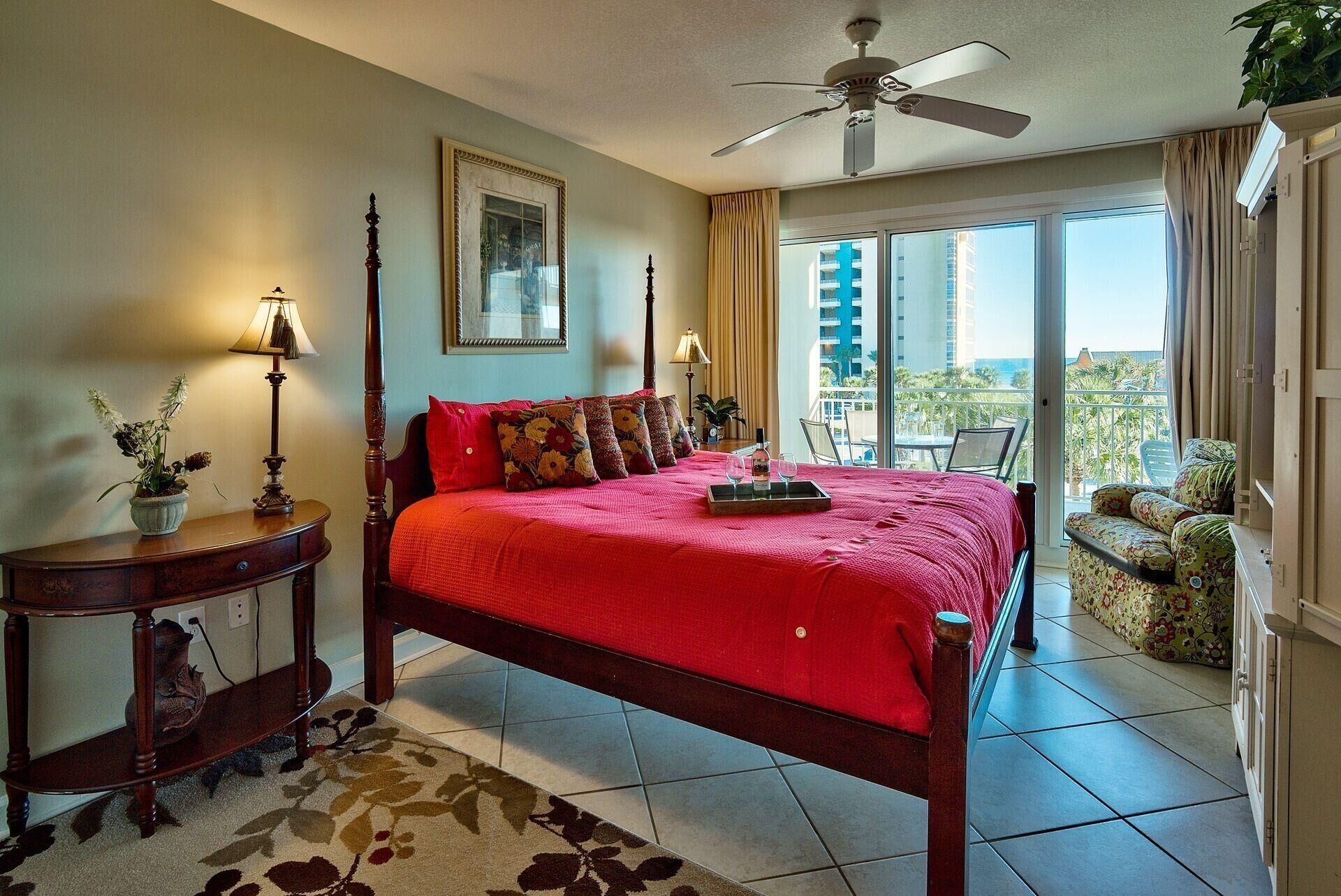 A hotel room with a large bed, a ceiling fan, a balcony with a view, and a small table with a lamp.
