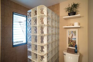A bathroom with a glass block wall, a toilet, a window with blinds, a shelf with a plant, and a framed picture.