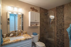 A bathroom with a granite countertop sink, a white toilet, a glass shower enclosure, and a mirror with lights.