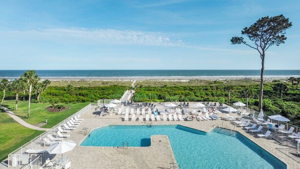 Ein Poolbereich mit Liegestühlen und Sonnenschirmen, mit Blick auf den Strand und einem großen Baum.
