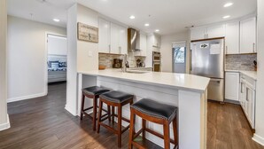 Una cocina moderna con muebles blancos, una isla central con taburetes de bar, un refrigerador de acero inoxidable y un protector contra salpicaduras con un letrero de temática playera.