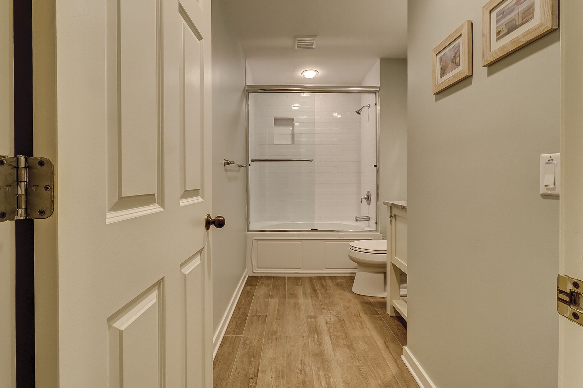 Un couloir étroit mène à une salle de bain équipée d’une douche, de toilettes et ornée de tableaux encadrés au mur.